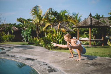 Aktif giyimli esnek, genç, çıplak ayaklı kadın elinde denge kurarken tropik bir tatil köyünde yüzme havuzunun yanında Firefly yoga pozu veriyor.