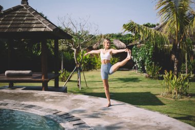 Güneşli bir günde yeşil parkta güneşli bir günde Utthita Hasta Padangusthasana yogası yaparken havuz kenarında durup kollarını esneten zayıf bir kadının bütün vücut görüntüsü.