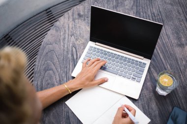 Yukarıdan gelen isimsiz kadın serbest yazar elinde bir bardak limon suyu akıllı telefonuyla ahşap masada oturuyor ve ekrana bakarken dizüstü bilgisayardan notlar alıyor ve gün ışığında kafede işe başlamak için çalışıyor.