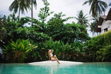 Yüzme havuzunun kenarında, güpegündüz tropik yeşil bitkilerin yanında oturan ve tatil yerlerinde yaz tatilinin tadını çıkaran genç bir kadının yan görüntüsü.