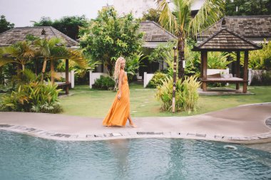 Havuz kenarında yaz tatilinde egzotik bitkilerle birlikte havuz kenarında yürüyen şık giyinmiş, yalınayak, genç ve güzel bir kadının yan görüntüsü.