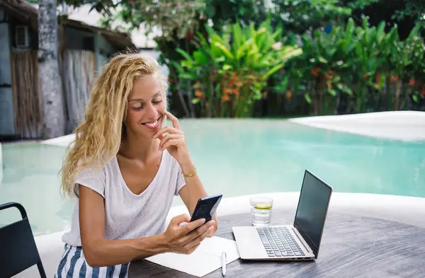 Smiling female freelancer in casual clothes sitting at table in resort with laptop and texting on mobile phone while working on remote project