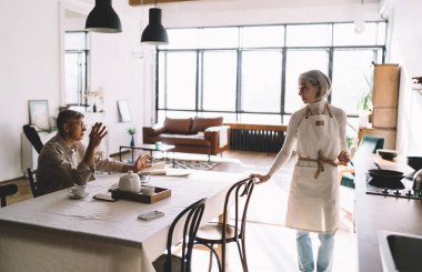 Masada dizüstü bilgisayarla oturan ve önlük giymiş yaşlı bir kadınla konuşan ve birlikte mutfakta boş vakit geçiren yaşlı bir erkeğin yan görüntüsü.