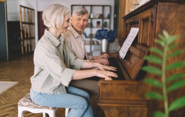 Olumlu yaşlı karı-koca gündelik kıyafetleriyle piyano çalarken evde birlikte vakit geçirirken müzik kağıtlarıyla melodi çalıyorlar.