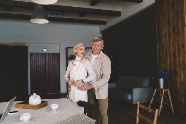 Önlüklü, gülümseyen orta yaşlı bir kadın romantik kocaya bakıyor. Evdeki mutfağın yanında durmuş, onu kucaklıyor.