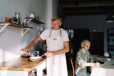 Mutfak tezgahında, karısının yanında nefis bir kahvaltı hazırlayan önlüklü olgun bir adam hafta sonu boyunca evdeki masada deftere bir şeyler yazıyor.