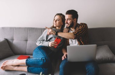 İyimser sakallı bir adam gülümseyerek ve parmaklarıyla işaret ederek kız arkadaşıyla rahat bir kanepede oturur ve başka tarafa bakar.