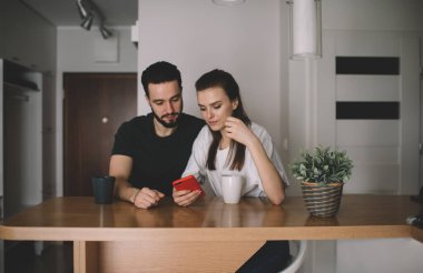 Konsantre olmuş genç adam ve kadın gündelik giysiler içinde masada oturuyor ve modern oturma odasında internette gezinirken cep telefonlarını karıştırıyor.