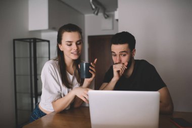 Siyah tişörtlü sakallı adam bilgisayardan genç kız arkadaşıyla şok edici videolar izlerken ağızını eliyle kapatıyor.