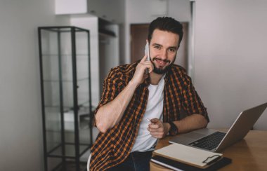 Gündelik kıyafetli genç, pozitif sakallı bir yönetici. Masada dizüstü bilgisayarla oturuyor ve iş arkadaşıyla cep telefonuyla konuşurken kameraya bakıyor.