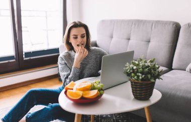 Odaklanmış genç bir kadın internette gezinmek ve meyve yemek için dizüstü bilgisayar kullanırken gri renkli parlak, rahat bir odada çalışıyor.