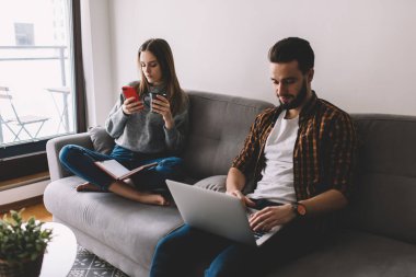 Erkek bilişim uzmanı, uzaktan programlama için bilgisayarını kullanırken, kahve fincanı olan kız arkadaşı akıllı telefondan sosyal medyada geziniyor.