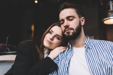Koyu renk saçlı, şık elbiseli, sevgi dolu genç bir çift romantik bir buluşmada sarılıp, gözlerini kaçırıyor.