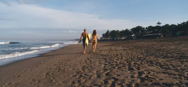 Sarı sörf tahtası taşıyan bir adam ve güneşli bir günde kumlu sahilde bikinili bir kadın.