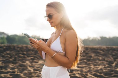 Uzun saçlı, güneş ışığına karşı duran ve tatillerde cep telefonuyla mesajlaşan bikinili genç bir kadın.