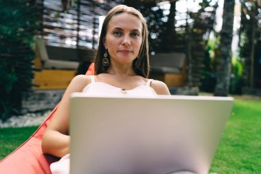 Yeşil çimlerde oturan ve parktaki kameraya bakarken dizüstü bilgisayarda çalışan serbest çalışan genç bir kadın.