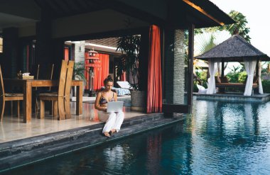 Açık yüzme havuzuna karşı terasta oturan ve tropik otellerde uzaktan görev yapan kadın serbest çalışan.