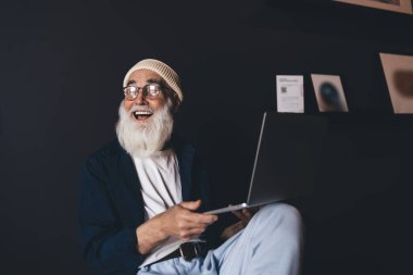 Gözlüklü, siyah duvarlı ve fotoğraf çerçeveli bir odada dizüstü bilgisayarlı bir sandalyede otururken yan yan yan yan bakan pozitif yaşlı bir erkek.