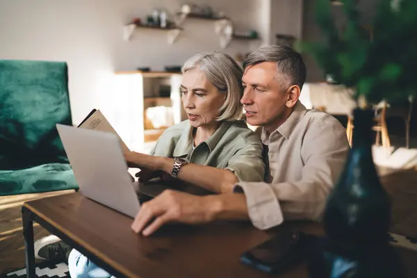 Focused aged couple with gray hair and in casual outfits sitting at wooden coffee table woman pointing at information in book while using laptop