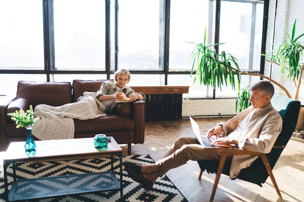 stock image Concentrated mature man sitting in armchair using laptop while woman reading book on comfortable sofa in living room at home