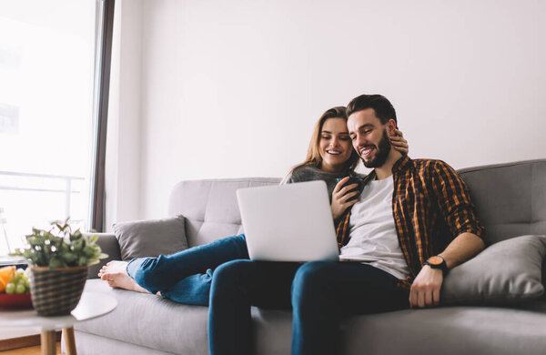 Cheerful couple in casual wear smiling and watching movie on laptop while resting on comfortable sofa and hugging at home