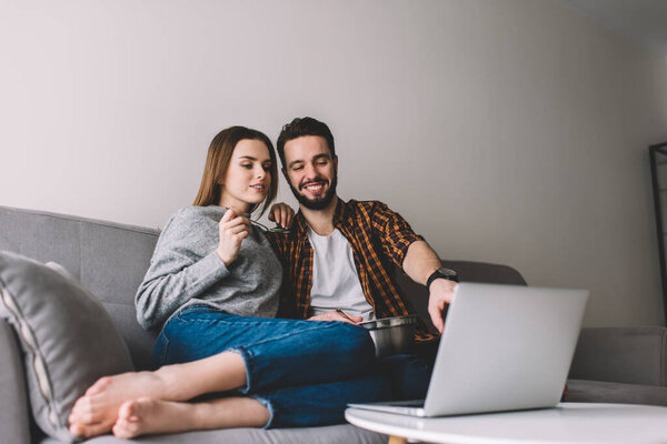 Carefree boyfriend and girlfriend resting at comfortable sofa spending together weekend for watching video on laptop