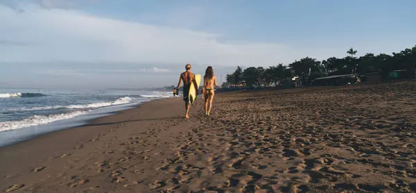 Back view full body of man carrying yellow surfboard and woman in bikini strolling along sandy seashore on sunny day