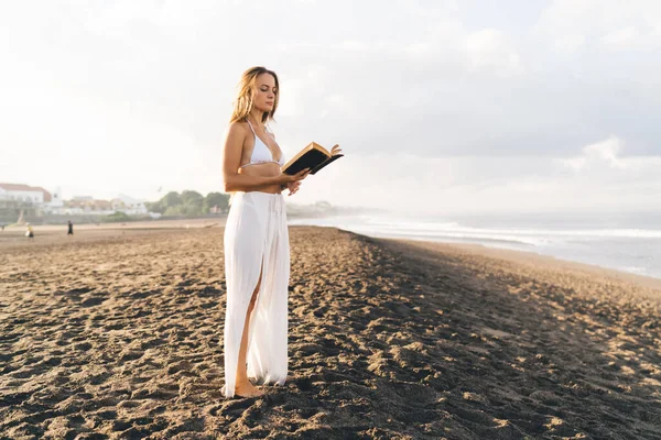 Full body of female in white bikini with long skirt standing on sandy seashore and reading interesting novel during summer weekend