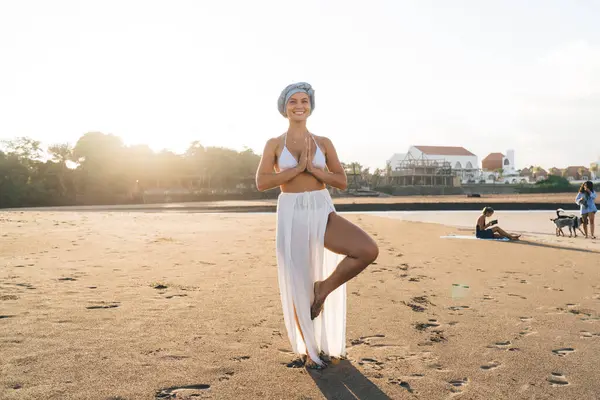 Sarıklı ve beyaz mayo giymiş sakin bir kadın Vrikshasana 'da kumlu sahilde yoga yapıyor.