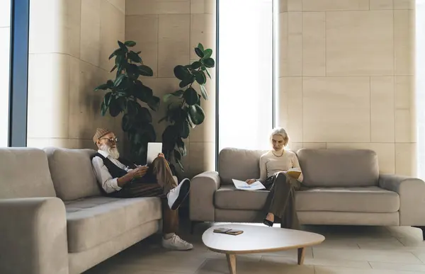 Side view of elderly male with laptop and looking at screen while female busy with diagrams reading book and relaxing together on sofas near table with notepad in daylight illuminated room in office