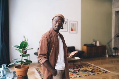 Ciddi bir Afro-Amerikalı erkek, elinde saksı bitkisiyle yerde duruyor ve evde gündüz vakti akıllı telefon kullanırken kafasını çeviriyor.