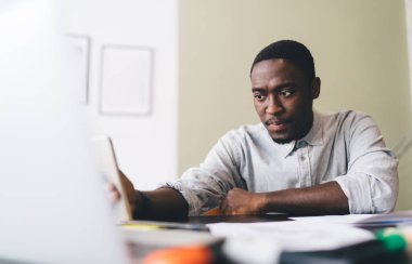 Ciddi bir Afrikalı Amerikalı erkek serbest çalışan gündelik giysiler içinde masada oturuyor ve internet üzerinden internet üzerinden proje üzerinde çalışıyor.