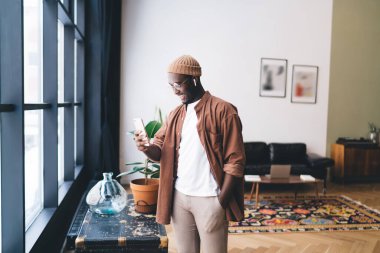 Afrika kökenli Amerikalı erkek başlıklı ve TWS kulaklıklı adam gün ışığında pencerenin yanında duruyor ve evde ekrana bakarken müzik listesini değiştiriyor.