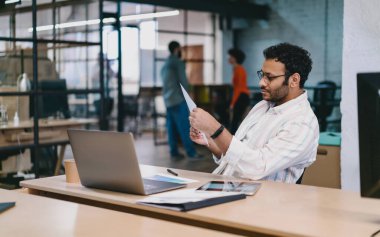 Sakallı, kıvırcık saçlı, zeki, rahat gömlekli, dizüstü bilgisayarlı ve yaratıcı çalışma alanında çalışırken elinde tuttuğu kağıtları okuyan profesyonel bir erkek.