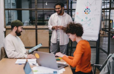 Bir grup çok ırklı iş arkadaşı, gündelik giysiler içinde kağıt tablet ve dizüstü bilgisayarlarla masada toplanıyor ve modern ofiste birlikte çalışırken projeyi tartışıyorlar.