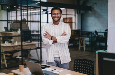 Modern çalışma ortamında gülümseyen ve kameraya bakarken elleri katlanmış, dizüstü bilgisayarlı ve bir fincan kahveli sıradan giyinen genç, pozitif sakallı bir erkek işçi.