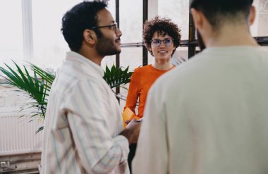 Gündelik giysiler ve gözlük takmış bir grup pozitif erkek ve kadın meslektaşlar modern ofiste ayakta dururken proje üzerinde tartışıyorlar.