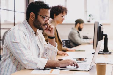 Ofis odasındaki dizüstü bilgisayarında iş arkadaşlarıyla çalışırken ahşap masada elleri çenesinde oturan yoğun sakallı, kıvırcık saçlı İspanyol erkek işçinin yan görüntüsü.