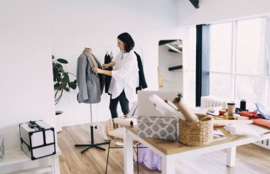 Stüdyoda yeni tasarım projesi üzerinde çalışırken, günlük kıyafetlerle manken sandığını ceketle ölçen kadın terzinin yan görüntüsü.