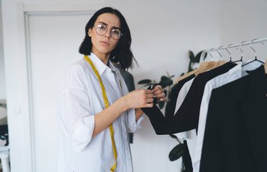 Gözlüklü, kendine güvenen genç bir kadın terzi ve kameraya bakan bir mezura ile askılık giymiş ve elinde siyah bir palto tutan bir sürü adamın yanında duruyor.