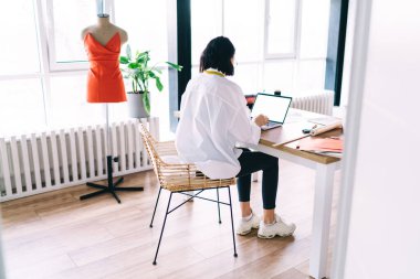 Beyaz tişörtlü, isimsiz bir kadının atölyede mankenlerle çalışırken laptopunu boş ekranla kullanışının tüm vücut arkası görüntüsü