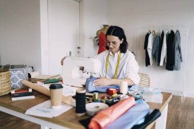 Boyunda mezurası olan hoş bir kadın terzi atölyede kumaşla çalışırken dikiş makinesiyle masada oturuyor.