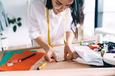 Gözlüklü ve beyaz gömlekli konsantre kadın tasarımcı masa üzerinde yeni tasarım projesi yaratırken mezurayla çalışıyor.