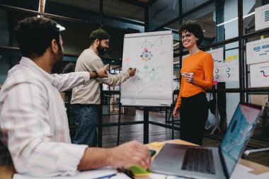 Neşeli erkek ve bayan meslektaşlar, modern çalışma ortamında çalışırken dizüstü bilgisayarla masada oturan erkek işçiyle birlikte projenin detaylarını incelerken,