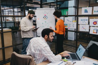 Çeşitli iş arkadaşları gündelik giysiler içinde kağıt pano ve çizelgelerle ayakta duruyorlar ve modern ofiste proje üzerinde çalışırken dizüstü bilgisayar ve belgelerle masaya oturuyorlar.