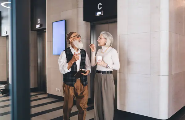 Confident elderly businesspeople in trendy clothes standing together in office hallway and discussing business strategy during coffee break while looking at each other