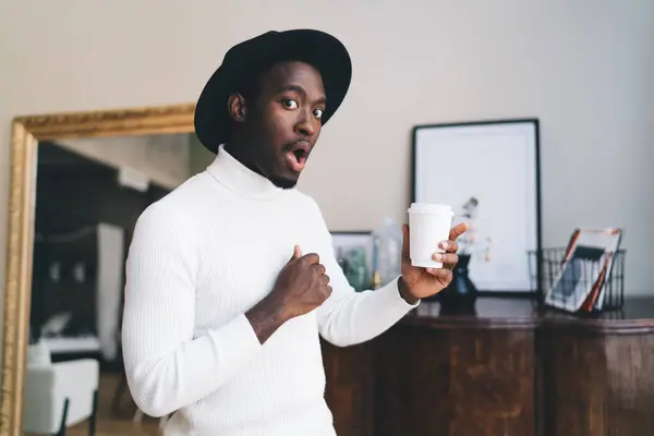 Side view of astonished African American male in black hat standing with cup of coffee in hand and pointing finger up while looking at camera near mirror with blurred reflection
