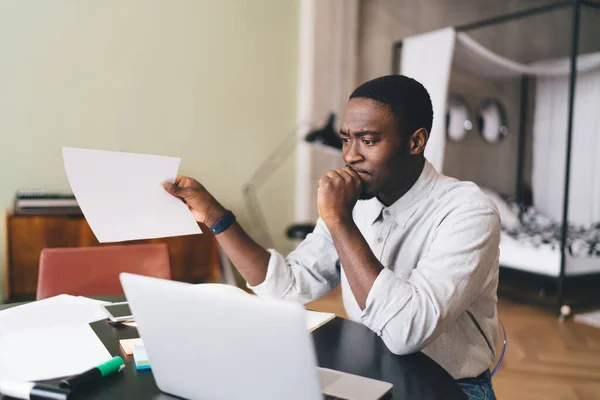 Troubled African American male freelancer sitting with fist touching lips at table with laptop documents while holding paper in hand and looking away in blurred living room
