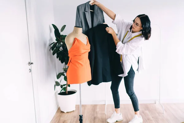 Full body of young female stylist in white shirt and glasses choosing jacket for stylish top on mannequin while working in studio
