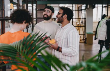 Modern ofis koridorunda dikilip palmiye yapraklarıyla cam duvarda tartışan gündelik giysiler içinde pozitif etnik çeşitliliğe sahip bir grup erkek ve kadın meslektaşız.